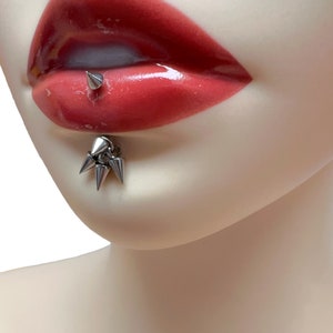 May include: A silver lip piercing with multiple spikes. The piercing is in a model's lower lip.