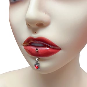 May include: A silver lip piercing with a dangling red gem. The gem is set in a square-shaped frame.