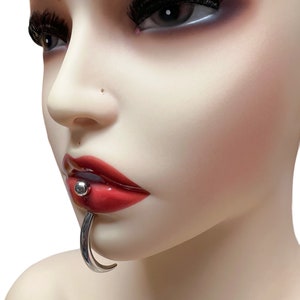 May include: A silver lip ring with a small ball on the end. The ring is curved and goes through the lip. The model is wearing red lipstick.