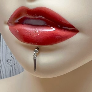 Może przedstawiać: Srebrny piercing do ust z małą kulką na górze i ostrym kolcem zwisającym w dół.