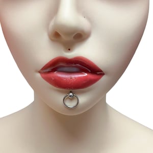 Può includere: Un anello per labbra argentato con una piccola palla sulla parte superiore. L'anello è nel labbro inferiore di una persona.