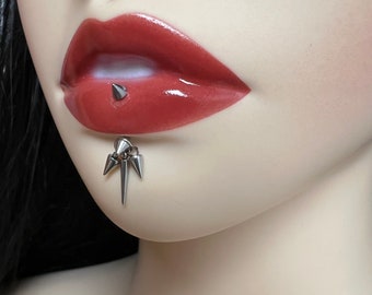 Può includere: Primo piano della parte inferiore del viso di una persona con un piercing al labbro argentato. Il piercing presenta un piccolo perno appuntito sul labbro inferiore e un design più grande a più punte sul mento.