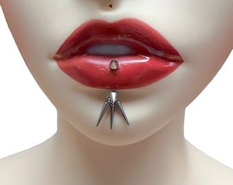 May include: A silver lip piercing with three sharp points. The piercing is in a person's lower lip.