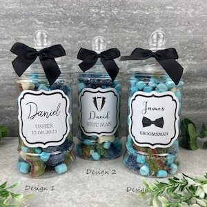 Può includere: Tre barattoli di vetro trasparente con coperchi neri e fiocchi neri pieni di caramelle blu e bianche. Ogni barattolo ha un'etichetta bianca con testo nero. Le etichette recitano "Daniel Usher 17.08.2023", "David Best Man" e "James Groomman".