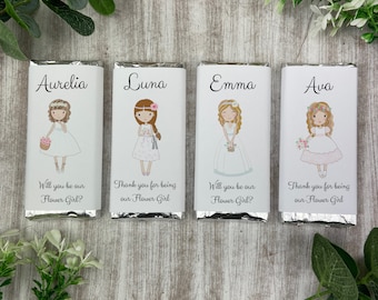 May include: Four personalized chocolate bar wrappers with illustrations of girls in flower girl dresses. The wrappers have the names Aurelia, Luna, Emma, and Ava printed on them. Each wrapper has a different message, such as "Will you be our Flower Girl?" or "Thank you for being our Flower Girl."