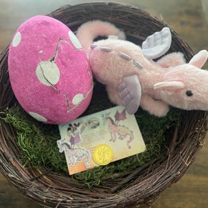 May include: A pink dragon plush toy with purple wings is nestled in a wicker basket with a pink and white speckled egg and a card with a dragon illustration.
