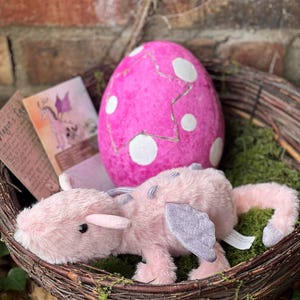 May include: A pink plush dragon toy with gray wings and accents rests in a woven basket with a pink egg. The egg has white spots and a cracked design. A card with a dragon illustration is also in the basket.