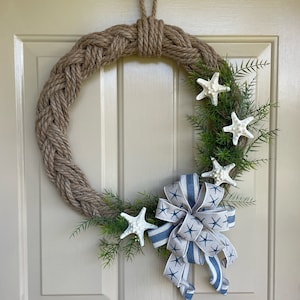 May include: A natural rope wreath with a blue and white striped bow and white starfish accents. The wreath is decorated with green foliage.