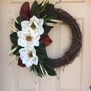 May include: A brown grapevine wreath with white magnolia flowers and green leaves. The wreath is decorated with small white sprigs.