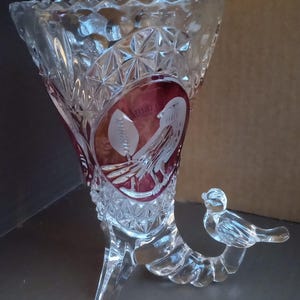 May include: Clear crystal vase with a flared top and three legs. The vase features a red oval design with a bird and leaf etching. A small bird figure is perched on a curved support.