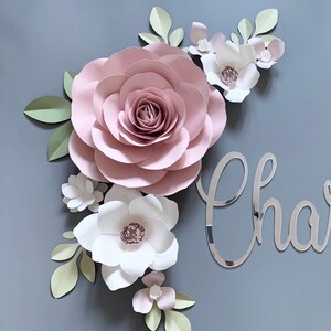 May include: A paper flower arrangement with a large pink rose, smaller white and pink flowers, and green leaves. The name "Charlie" is written in silver mirror acrylic letters.