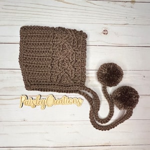 Op de afbeelding: Een bruine gehaakte baby muts met twee bruine pompoenen aan de bandjes. De muts heeft een kabelbreipatroon. De tekst "Paisley Creations" is zichtbaar op een houten bord.