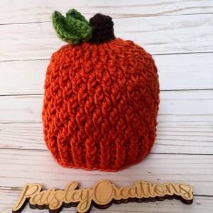 Könnte beinhalten: Ein handgefertigter orangefarbener Häkel-Kürbishut mit einem grünen Blatt und einem braunen Stiel. Der Hut besteht aus strukturiertem Garn und liegt auf einer hellen Holzoberfläche. Der Hut ist ein Herbstartikel.