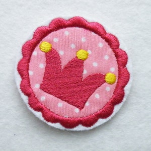 Könnte beinhalten: Gestickter Aufnäher mit einem rosa Kronen-Design und gelben Akzenten. Die Krone befindet sich auf einem rosa Stoff mit weißen Punkten, umgeben von einem rosa, gewellten Rand und einem weißen Rand. Geeignet für Kleidung oder Accessoires.