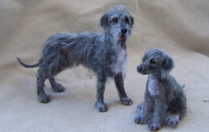 May include: Two needle felted grey Irish Wolfhounds. The larger dog is standing with its tail curled. The smaller dog is sitting with its ears perked up.