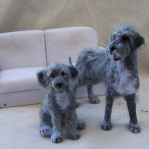May include: Two gray felt dog figurines, one larger than the other, sitting in front of a white couch. The larger dog is standing with its head tilted to the side, while the smaller dog is sitting with its paws crossed.