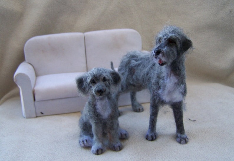 May include: Two gray felt dog figurines, one larger than the other, sitting in front of a white couch. The larger dog is standing with its head tilted to the side, while the smaller dog is sitting with its paws crossed.