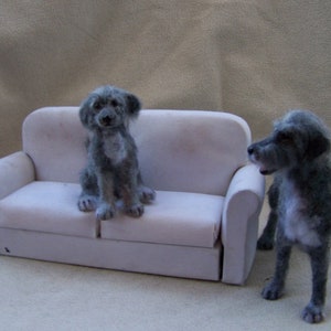 May include: Two gray, furry dogs sitting on a white couch. One dog is sitting on the couch, while the other dog is standing next to the couch.