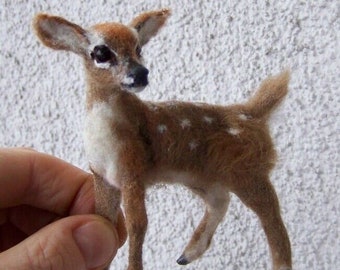 May include: A small, brown and white needle felted fawn with white spots. The fawn is standing on a white textured surface.
