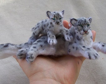 May include: Two felted snow leopard sculptures, one adult and one cub, are posed on a hand. The adult snow leopard is lying down with its head turned to the side, while the cub is sitting on top of the adult's back. Both sculptures are made of white wool with black spots.