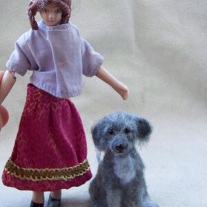 May include: A porcelain doll wearing a purple shirt and a burgundy skirt with gold trim stands next to a gray felt dog.