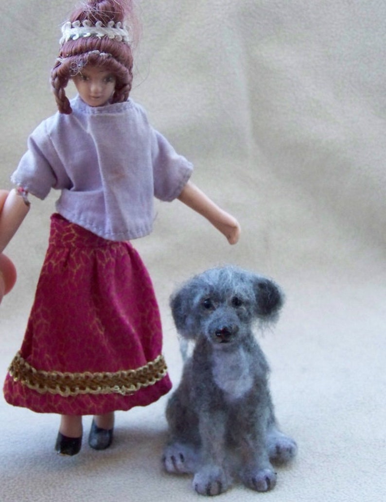 May include: A porcelain doll wearing a purple shirt and a burgundy skirt with gold trim stands next to a gray felt dog.