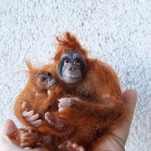 May include: A close-up of a pair of orangutan dolls, one adult and one baby, made of soft, reddish-brown fur. The adult orangutan is holding the baby orangutan in its arms.