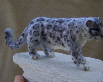 May include: A white and gray snow leopard figurine with black spots, standing on a white rock. The figurine is made of wool felt and is very realistic.