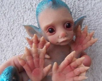 Puede incluir: Una pequeña muñeca hecha a mano, de color azul y rosa, con un mohicano azul puntiagudo, ojos grandes y manos palmeadas. La muñeca está hecha de un material suave y realista y tiene un aspecto caprichoso y de hada.
