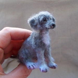 May include: A small, gray and white needle felted dog figurine. The dog is sitting with its head tilted to the side and its eyes closed.