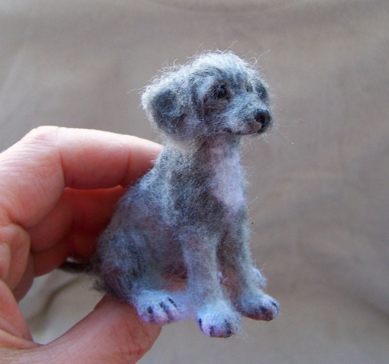 May include: A small, gray and white needle felted dog figurine. The dog is sitting with its head tilted to the side and its eyes closed.