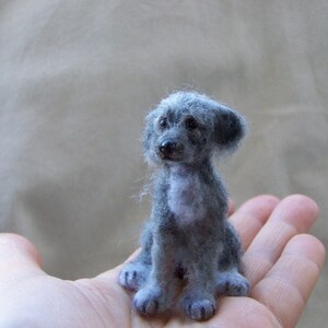 May include: A small, gray, needle felted dog with a white patch on its chest. The dog is sitting on a person's hand.