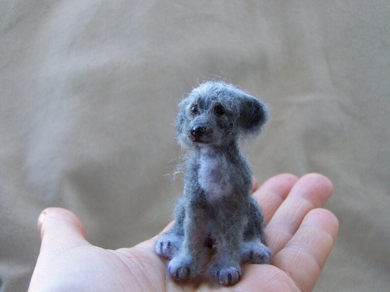 May include: A small, gray, needle felted dog with a white patch on its chest. The dog is sitting on a person's hand.