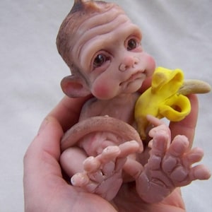 May include: A small, realistic-looking monkey doll with brown fur, holding a yellow banana. The doll has a detailed face with brown eyes and a pink blush.