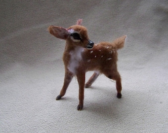 May include: A small, brown and white fawn with white spots, made of felt. The fawn is standing on a light brown surface.