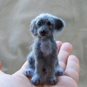 May include: A small, gray, felted dog figurine with a fluffy coat. The figurine is sitting on a person's hand.