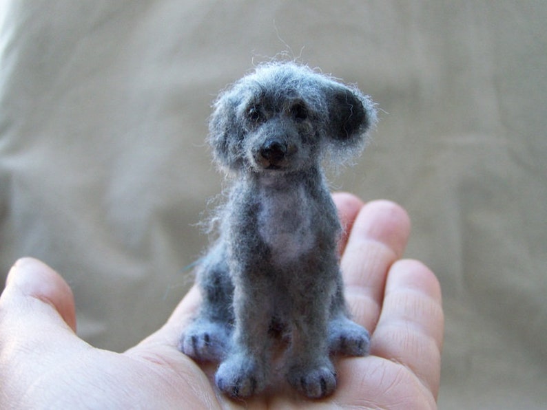May include: A small, gray, felted dog figurine with a fluffy coat. The figurine is sitting on a person's hand.