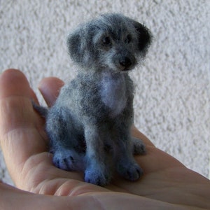 May include: A small, gray, needle-felted dog figurine sitting on a hand. The dog has a fluffy coat and is sitting with its paws tucked under its body.