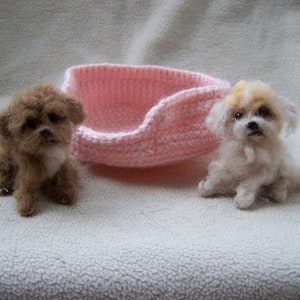 May include: Two small, felted dogs, one brown and one white, sitting on a white fluffy surface. A pink crocheted dog bed is in the background.
