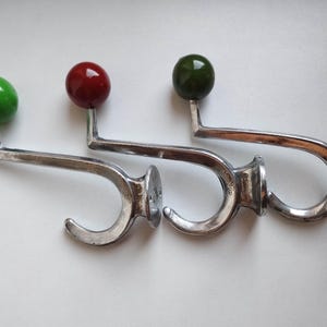 May include: Three metal wall hooks with a curved silver-toned finish and a spherical knob at the top. Each hook features a different colored knob: one bright green, one deep red, and one dark olive green. The hooks have a circular base for mounting.