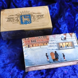May include: Two vintage decorative boxes. The larger box has a cream-colored lid with a crest and the word GOTLAND. The smaller box features a winter scene with people and a building. Both boxes are on a blue velvet surface.