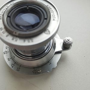 Puede incluir: Un objetivo de c&aacute;mara plateado vintage con componentes internos negros. La lente tiene una serie de n&uacute;meros y marcas, incluyendo "ИНДУСТАР-50" y "N66001204". Se ve un peque&ntilde;o bot&oacute;n texturizado en el lateral.