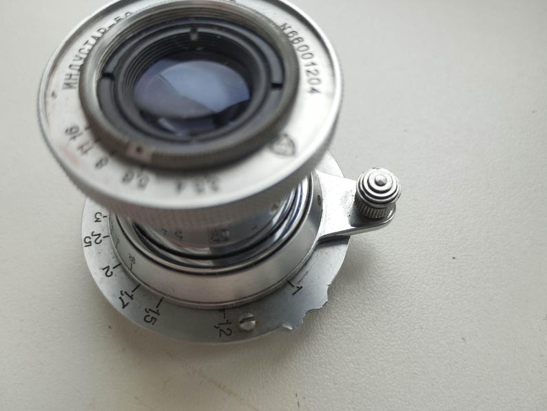 Puede incluir: Un objetivo de c&aacute;mara plateado vintage con componentes internos negros. La lente tiene una serie de n&uacute;meros y marcas, incluyendo "ИНДУСТАР-50" y "N66001204". Se ve un peque&ntilde;o bot&oacute;n texturizado en el lateral.