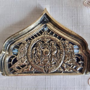 May include: Ornate, gold-toned religious icon with a central circular relief depicting figures, possibly saints or biblical characters. The icon has a dark blue and light blue background, and is framed by a decorative border. The top is pointed.