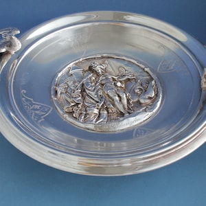 May include: A silver serving dish with two bird handles and a central embossed scene of a woman holding a staff and a bird. The scene is surrounded by a decorative border and the word "Valkyria" is inscribed below.