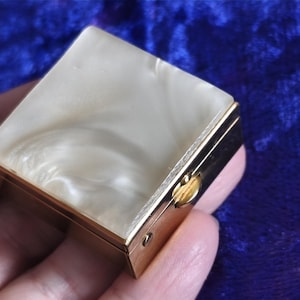 May include: A small, square, gold-toned pillbox with a pearlescent, off-white lid. The lid has a swirling pattern and a gold-colored clasp. The box is held in a hand against a dark blue background.