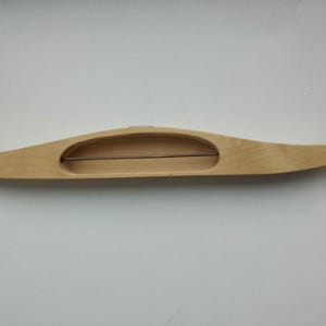 Peut inclure: Une navette de tissage en bois clair, de forme incurvée, avec une ouverture centrale. Une fine tige métallique traverse le centre de la navette. La navette est conçue pour le tissage.