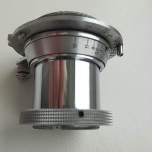 Puede incluir: Primer plano de una lente de c&aacute;mara plateada vintage. La lente tiene un anillo de enfoque estriado, ajustes numerados y un peque&ntilde;o bot&oacute;n de ajuste. La lente cil&iacute;ndrica tiene una superficie reflectante.