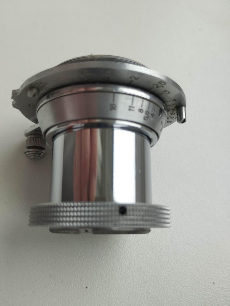 Puede incluir: Primer plano de una lente de c&aacute;mara plateada vintage. La lente tiene un anillo de enfoque estriado, ajustes numerados y un peque&ntilde;o bot&oacute;n de ajuste. La lente cil&iacute;ndrica tiene una superficie reflectante.