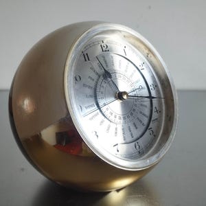 May include: A spherical desk clock with a polished gold-tone metal casing and a silver-toned face. The clock features black Arabic numerals, thin black hands, and an inner rotating dial displaying various global city names and a 24-hour scale.